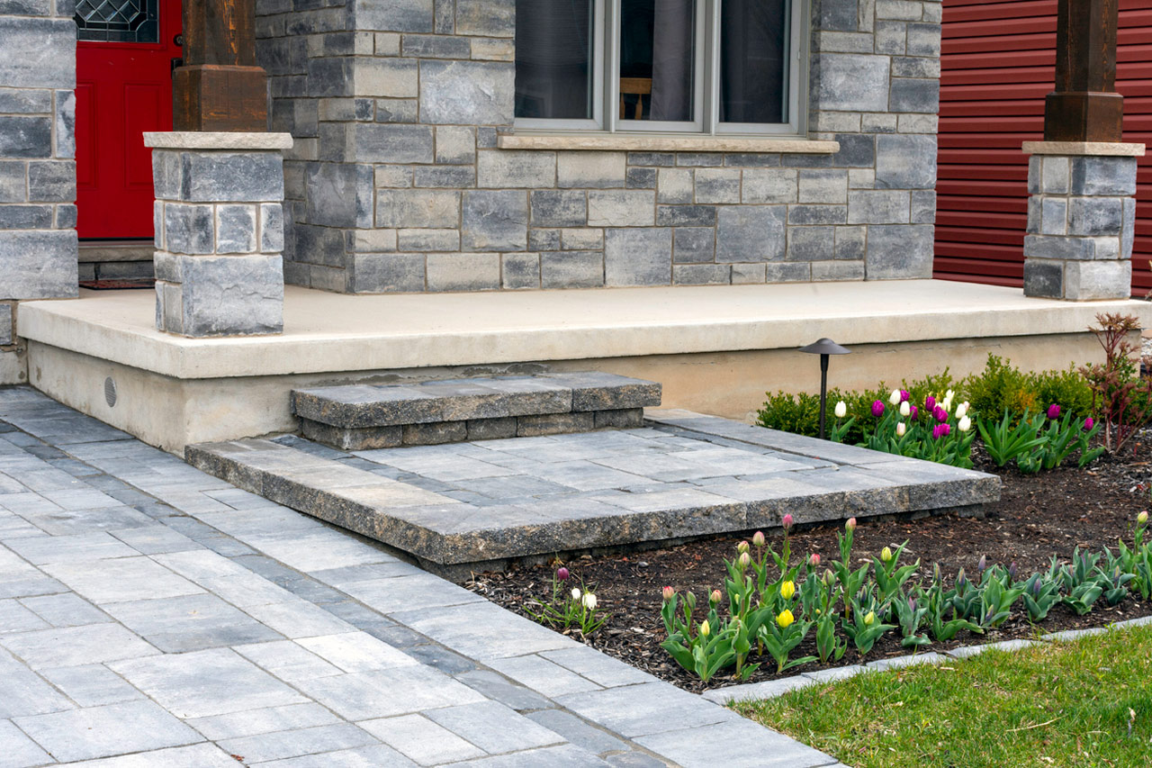 A clean, modern front porch featuring a smooth concrete landing with stone steps leading up to a red door. The entry is framed by gray stone pillars and flanked by landscaped flower beds with colorful tulips, all connected by a paved walkway for a welcoming and elegant approach.
