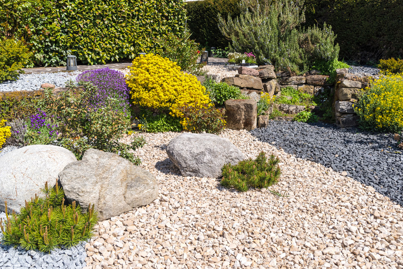 A precisely built angled retaining wall made of textured gray stone blocks, forming a corner that includes a built-in bench and garden bed. The structure supports a sloped landscape with young plants and mulch, demonstrating functional and aesthetic hardscape design.