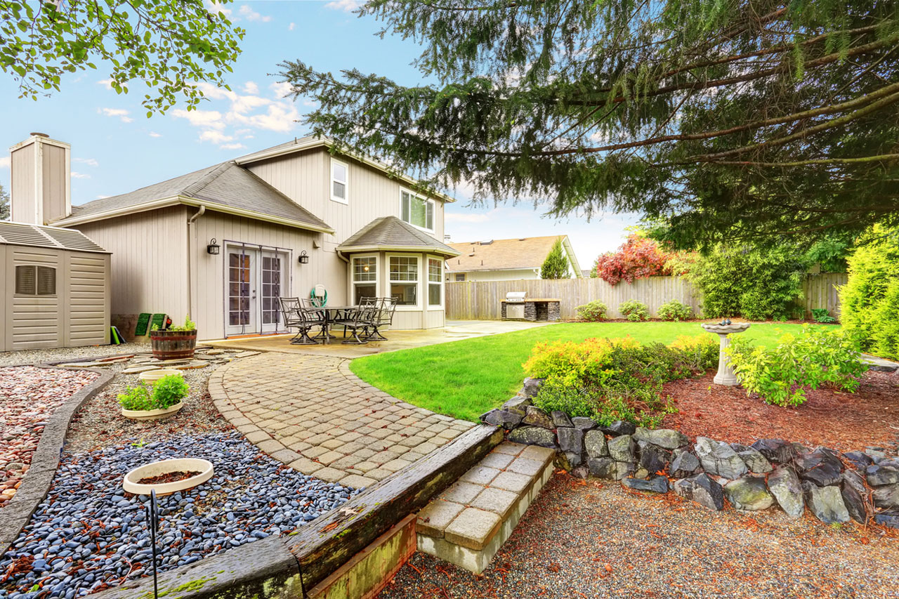 A well-designed backyard with a winding paver path leading through a mix of gravel, mulch, and rock gardens. The space includes a birdbath, flower beds, a patio area, and mature trees, offering a harmonious blend of hardscaping and natural beauty.