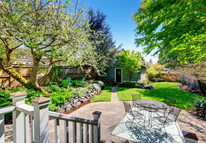 A beautifully landscaped backyard featuring a blooming white flowering tree, lush green lawn, and winding garden paths. The space includes stone retaining walls, colorful plantings, and a patio with a metal table and chairs on a patterned rug, framed by a white railing and mature trees under a bright blue sky.